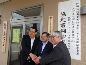 看板の前で市長、駄科区長、原社長握手